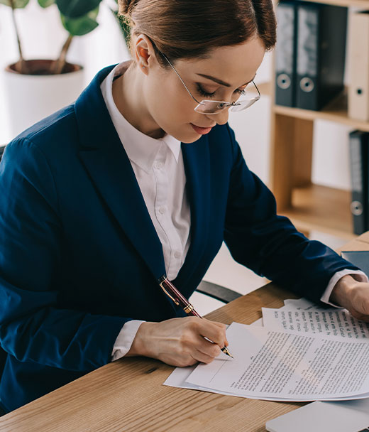 A woman in a blue blazer and glasses signing documents at a desk in an office setting